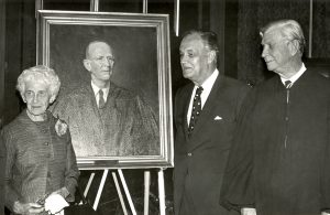 Photo of two men and a woman standing beside a portrait on an easel. 
