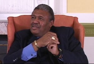 John Charles Thomas speaking during the August 2007 interview. He is seated in a chair wearing a suit and tie.