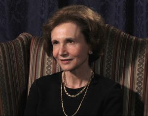 Barbara Milano Keenan speaking during the 2013 interview. She is seated in a chair, wearing a black blouse and gold jewelry.