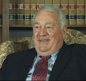 Lawrence L. Koontz speaking during the 2013 interview. He is seated in a chair in front of a book shelf wearing a suit and tie.