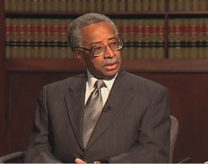 Hon. James W. Benton speaking during the March 2013 interview. He is seated in front of a book case wearing a suit and tie.