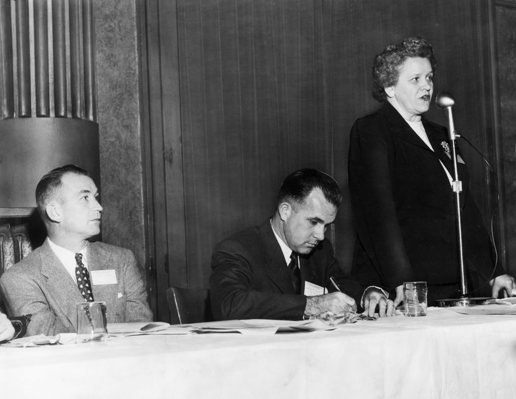 Hon. Odessa Pittard Bailey stands speaking at a microphone wearing a suit with a brooch. Two men are seated at the panel table beside her wearing suits and ties.