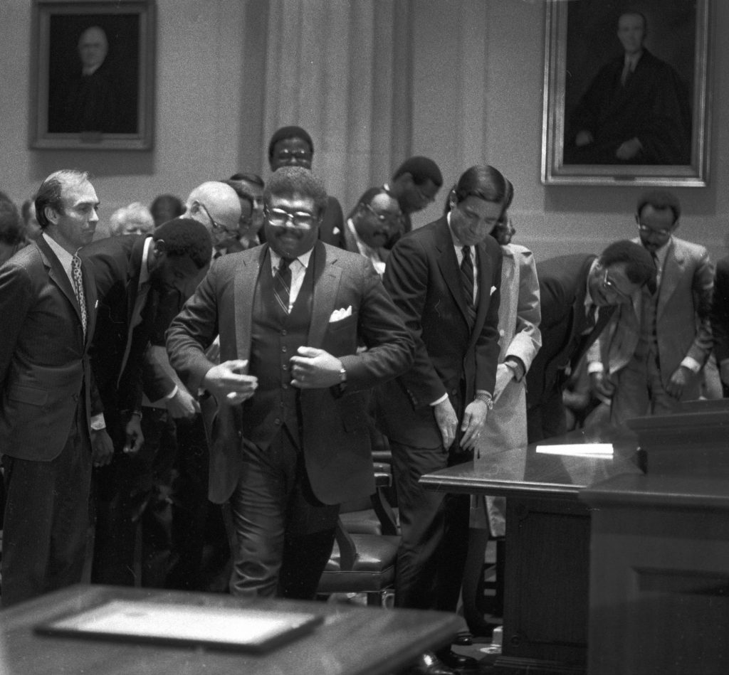 Hon. John Charles Thomas walking in the Supreme Court of Virginia courtroom surrounded by other men. He is wearing a three piece suit. 