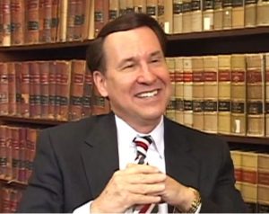 Robert N. Baldwin speaking during the 2010 interview. He is seated in a chair in front of a book shelf wearing a suit and tie.