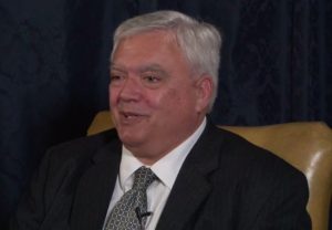 Jack Bruce speaking during the 2013 interview. He is seated in a chair wearing a suit and tie.