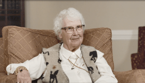 Jean Clements during the 2024 interview. She is seated in a large armchair, wearing a collared white shirt, soft tan and black vest, and glasses.