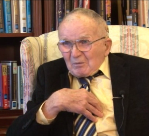 Henry Hudson Whiting speaking during the 2011 interview. He is seated in a chair in front of a book shelf wearing a suit and tie. 